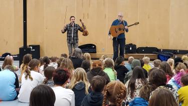 In an elementary school gymnasium full of students watching a performance by two men, one with a fiddle and one with a guitar. 