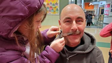 A student with a pair of scissors trimming a male teachers mustache for a fundraiser. 