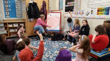 Students in a classroom sitting in a circle, teacher sitting hear flip chart with a giant heart on it.