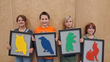 Four students standing in a row, each holding up a sign with the symbol of their house team on it (owl, raven, bear or coyote)