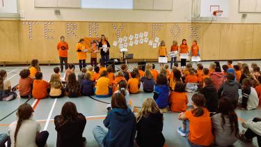 Elementary school students sitting in an assembly in the gymnasium, many students wearing orange shirts. 