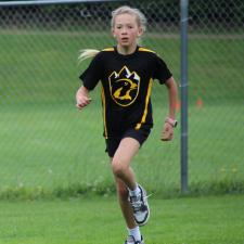 A blond girl in a black track uniform runs across a grassy field
