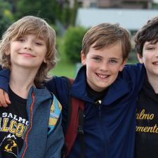 Three boys smile outdoors with their arms around one another's shoulders