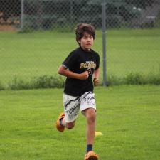 A child in a black shirt and light coloured shorts runs through a grassy field in a track competition