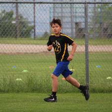 A child in a black shirt and blue shorts runs through a grassy field with a chainlink fence behind himin a track competition