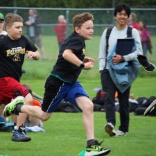 Two boys sprint to the finish while a smiling spectator looks on in a school track competition in a grass field